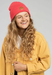 Różowy, prążkowany beret z okrągłym, jasnobrązowym skórzanym logo, noszony z żółtą dżinsową kurtką. Modelka ma długie, falowane włosy.