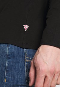 Czarna koszula z długim rękawem wykonana z bawełny, z małą naszywką z logo "GUESS" w kolorze czerwonym i białym na dolnym brzegu, w zestawie z niebieskimi dżinsami.