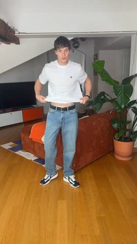 Bărbat tânăr, îmbrăcat în tricou alb și blugi albaștri, ridică ușor cămașa în sufragerie cu canapea portocalie și plantă mare în ghiveci.