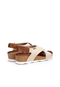 Beżowe sandały z krzyżowym paskiem, brązowe skórzane akcenty, korek na obcasie i biała podeszwa. Posiadają regulowany pasek w kostce z klamrą.