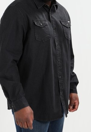Mann trägt ein schwarzes Hemd mit Knöpfen und Brusttaschen und blaue Jeans, steht vor einem schlichten hellen Hintergrund.