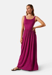 Robe maxi en fuchsia, sans manches avec des larges bretelles, taille froncée pour l'élasticité, jupe fluide et sandales avec des accents en perles.