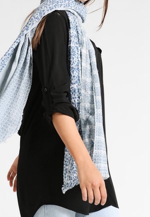 Femme portant un haut noir à manches longues, une écharpe bleu clair à motifs et un jean bleu clair, debout devant un fond blanc uni.