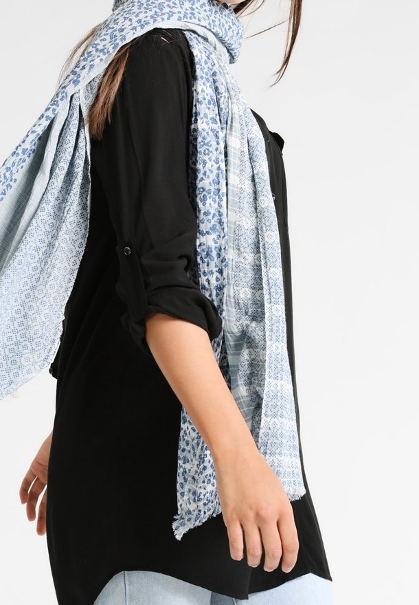 Femme portant un haut noir à manches longues, une écharpe bleu clair à motifs et un jean bleu clair, debout devant un fond blanc uni.