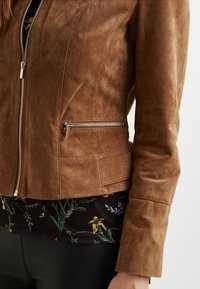 Chaqueta de ante marrón con diseño entallado, que cuenta con una cremallera frontal y bolsillos laterales con herrajes plateados. Superficie texturizada con detalles de costuras.