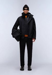 Czarna wodoodporna kurtka na pikowanej kamizelce, pomarańczowy golf, czarne spodnie i czarne buty. Akcesoria: czarna czapka i okulary przeciwsłoneczne, trzyma torbę.