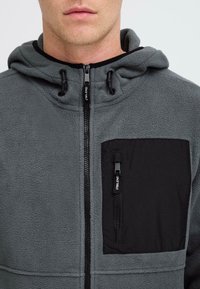 Mann trägt einen grauen Fleece-Hoodie mit einer schwarzen Reißverschlusstasche auf der Brust und schwarzen Reißverschluss-Schlaufen, zeigt Hals und unteres Gesicht.