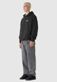 Svart hoodie med ficka fram och broderad logotyp, lager på lager över en grå tröja. Kombinerad med lösa grå jeans och svarta sneakers.