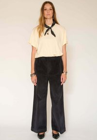 Chemise courte à manches courtes crème, pantalon large en velours côtelé noir et chaussures noires à motifs. Porte une écharpe noire et des bijoux en argent.