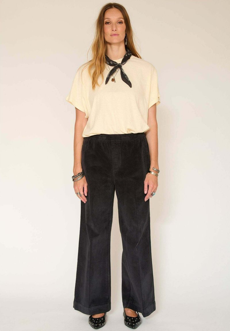 Chemise courte à manches courtes crème, pantalon large en velours côtelé noir et chaussures noires à motifs. Porte une écharpe noire et des bijoux en argent.