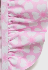 Vaalea vaaleanpunainen kangas, jossa on valkoisia sydämiä, ja siinä on röyhelöreuna sekä ommeltu nauha. Materiaali vaikuttaa pehmeältä ja sileältä.