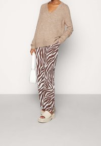 Beiger V-Ausschnitt-Pullover, braun-weiß gestreifte, weite Hose im Zebra-Look und klobige, cremefarbene Sandalen. Auch eine weiße Handtasche ist sichtbar.