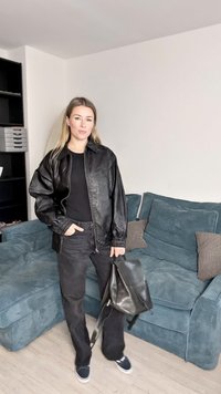 Svart jakke i imitert skinn med glidelås, ribbestrikkede mansjetter og en flat krage, kombinert med svarte bukser og en svart ryggsekk.