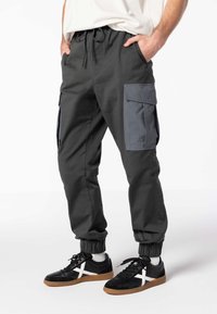 Pantaloni cargo in tessuto grigio scuro con polsini elasticizzati, dotati di due grandi tasche laterali di un grigio più chiaro. Abbinati a sneaker nere con dettagli bianchi.