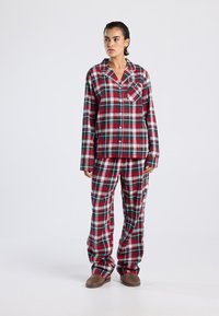 Woman standing wearing red, green, and white plaid long-sleeve button-up top and matching loose pants with brown slippers, neutral background.