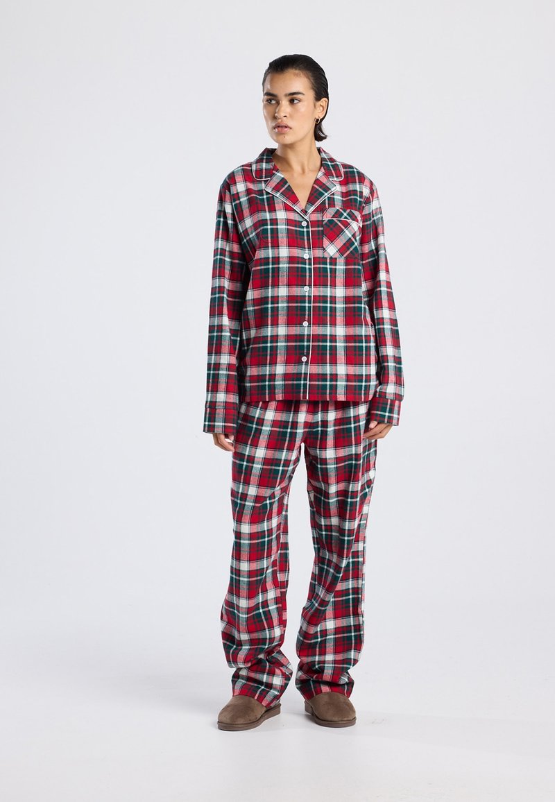 Woman standing wearing red, green, and white plaid long-sleeve button-up top and matching loose pants with brown slippers, neutral background.