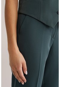 Close-up van de hand van een persoon die rust naast donkergroene op maat gemaakte broek en een bijpassend colbert met een zichtbare knoop.