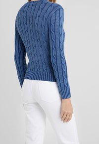 Kvinna i en tajt blå kabelstickad tröja och vita byxor, sedd bakifrån mot en enkel ljus bakgrund.