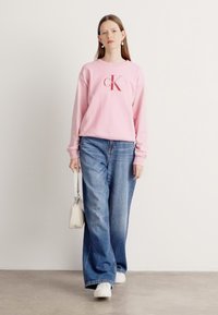 Felpa rosa con logo, abbinata a jeans blu larghi. Il modello tiene una piccola borsa bianca e indossa sneakers bianche. Abbigliamento semplice e casual.