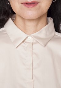 Partie inférieure du visage et cou d’une femme portant une chemise beige boutonnée avec col pointu et petites boucles d’oreilles anneaux.