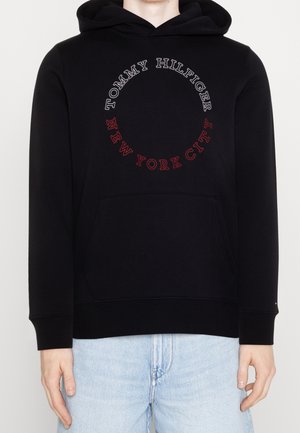 Osoba ubrana w czarną bluzę z kapturem z okrągłym napisem „Tommy Hilfiger” i „New York City” oraz jasnoniebieskie dżinsy, stojąca na jednolitym tle.