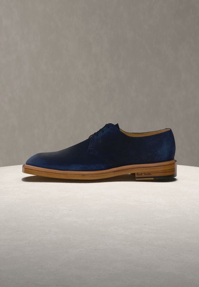Ανδρικό οξφορντ παπούτσι από μπλε ναυτικό σουέντ με ξύλινη σόλα και κορδόνια, εκτεθειμένο σε ανοιχτή επιφάνεια μπροστά σε ουδέτερο φόντο.