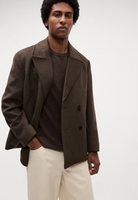 Bruine pea coat met grote revers en dubbele zwarte knopen. Draag deze over een gestructureerde bruine trui, gecombineerd met licht beige broek.