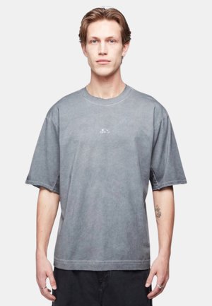 Jeune homme aux cheveux châtain clair portant un t-shirt gris délavé oversize et un pantalon noir, debout devant un fond blanc uni.