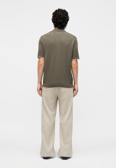 Άνδρας με σγουρά μαλλιά, φανερός από πίσω, φοράει ελαιδί κοντομάνικο πλεκτό πόλο και φαρδιά μπεζ παντελόνια, με λευκό φόντο.