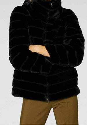 Personne portant une veste zippée en fausse fourrure noire avec des rayures texturées horizontales et un pantalon marron, bras croisés à la taille.