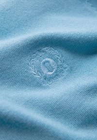 Tessuto blu chiaro con una consistenza morbida, caratterizzato da un logo ricamato al centro, circondato da un design floreale circolare.