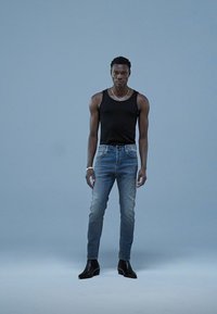 Svart ermeløs tanktopp, blå smale jeans med lett slitasje og svarte ankelstøvler. Modellen står foran en ensfarget lys blå bakgrunn.