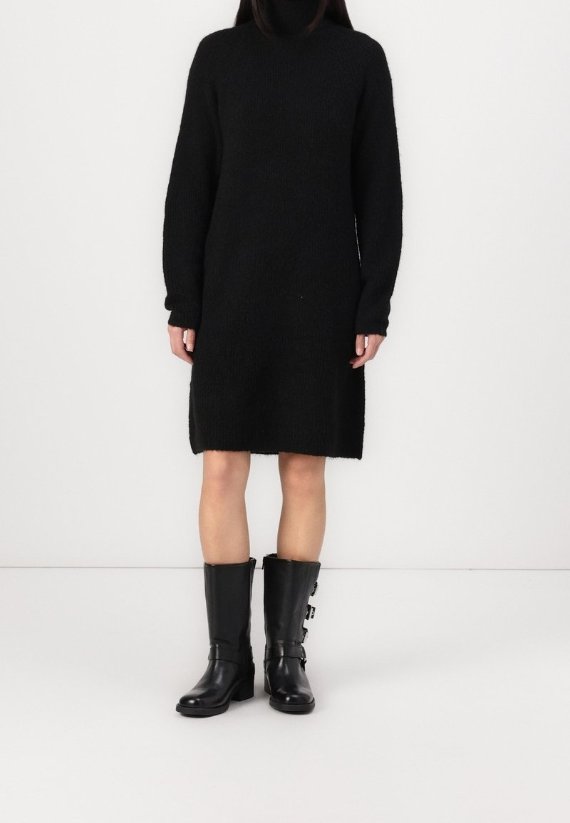 Femme portant une robe pull à col roulé noire longueur genou et des bottes en cuir noires mi-mollet avec des détails à boucle, debout devant un fond blanc.