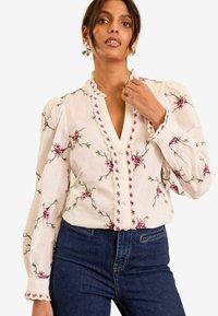 Blusa bianca ricamata con motivi floreali rosa, scollo a V ondulato, apertura con bottoni e maniche a sbuffo. Abbinata a jeans in denim scuro.