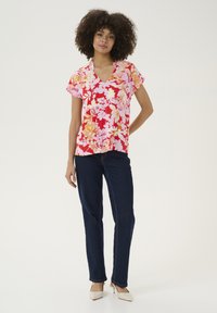 Cvetna majica s kratkimi rokavi v rdečih, roza in rumenih odtenkih. Oblika v vratu v obliki črke V, v kombinaciji z temno modrimi kavbojkami in bež petkami. Ozadje s ravno površino.