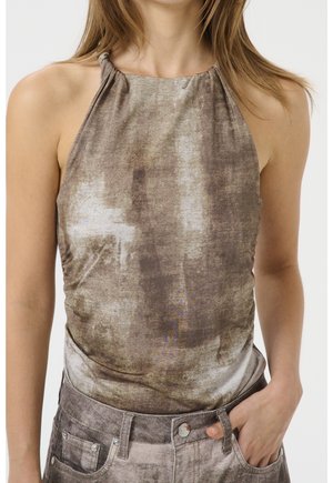 Frau trägt ärmelloses braun-beige Batik-Halfter-Top, das in passende hoch taillierte Jeans gesteckt ist, eine Hand in der Tasche.