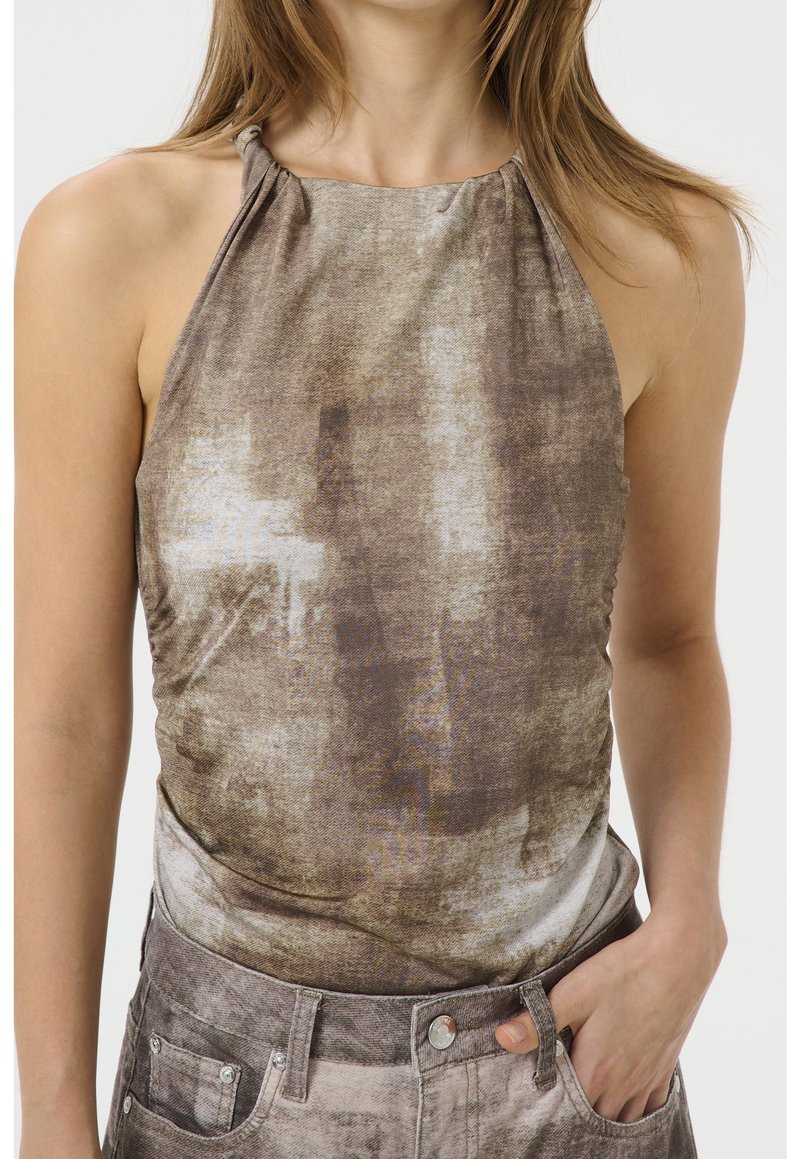 Femme portant un haut sans manches marron et beige tie-dye rentré dans un jean taille haute assorti, une main dans la poche.