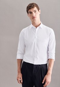 Weißes Hemd mit klassischem Kragen, langärmlig und bis zu den Ellenbogen gerollt, kombiniert mit schwarzen Hosen. Glattes Material, minimalistisches Design.