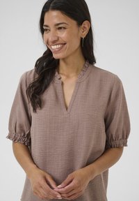 Blusa marrone, testurizzata, con scollatura a V e maniche corte a palloncino con polsini arricciati. Il tessuto presenta un motivo intrecciato sottile.