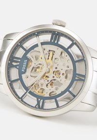 Montre-bracelet Fossil argentée avec cadran transparent montrant des engrenages dorés et argentés complexes et des chiffres romains en guise d'index horaires.
