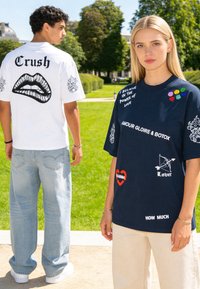 Due giovani adulti indossano magliette con scritte e simboli grafici; uno guarda di fronte, l’altro mostra la scritta "Crush" e un disegno di labbra su una maglietta bianca.
