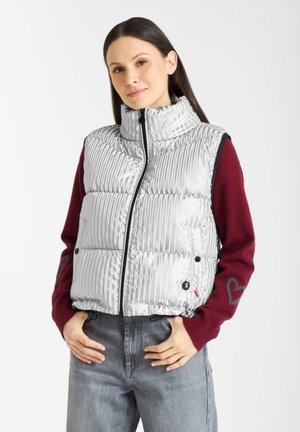 Zilveren puffer vest met verticale ribbels, hoge kraag, zwarte rits en zijzakken. Draagt over een bordeauxrode trui met hartdetail op de mouw.
