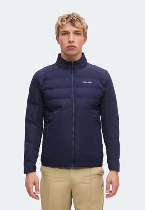 Junger Mann mit kurzen blonden Haaren trägt eine dunkelblaue Calvin Klein Steppjacke und beige Hose und steht vor einem einfachen hellen Hintergrund.
