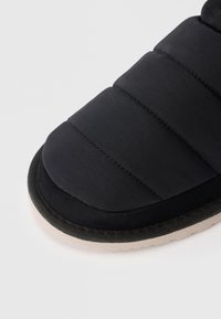 Pantofola nera imbottita con una parte superiore in tessuto liscio, punta rotonda e suola imbottita. Presenta una suola di colore più chiaro in contrasto.