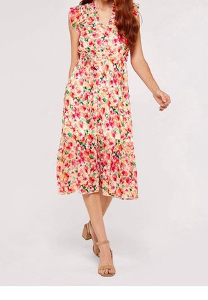 Kobieta ubrana w bezrękawową, kwiatową sukienkę midi z dekoltem w serek, falbaną na dole i paskiem w talii, zestawioną z beżowymi sandałami na obcasie typu klockowego z paskiem wokół kostki.