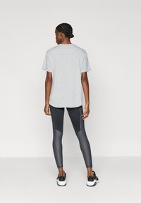 Graues Baumwoll-T-Shirt mit kurzen Ärmeln, kombiniert mit schwarzen Leggings mit strukturierten Einsätzen und glatter Oberfläche, getragen mit sportlichen schwarzen Turnschuhen.