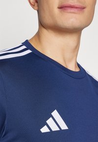 Marineblauw sportshirt met korte mouwen, ronde hals en drie witte strepen op de schouders. Bevat het witte merkembleem op de borst.