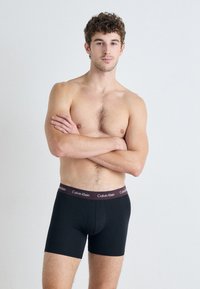 Čierne boxerky so stredným pásom, s jednofarebným dizajnom a pásom ozdobeným logom "Calvin Klein" v tmavo hnedej farbe.