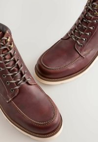 Botine din piele maro cu vârf rotund, accente cusute și șireturi gri. Talpă din cauciuc albă cu margine întunecată contrastantă.