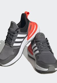 Zapatillas deportivas grises con parte superior de malla, franjas blancas a los lados y un acento naranja cerca del talón. Suelas de goma para mayor tracción.